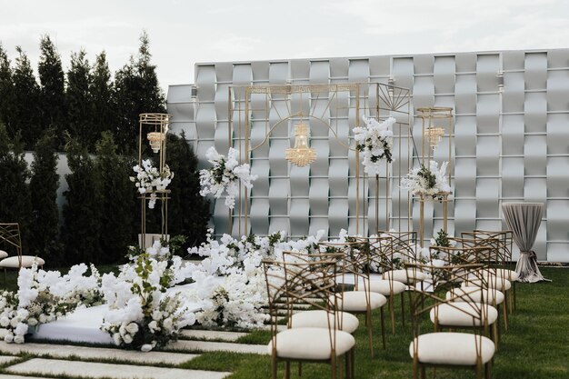 Une cérémonie de mariage dans un jardin avec des chaises blanches et un mur argenté avec des fleurs blanches.