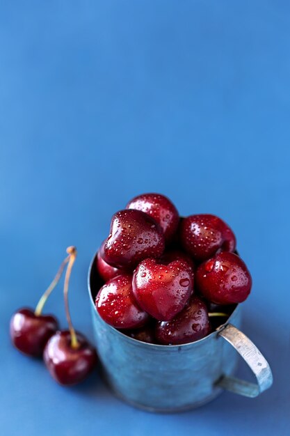 Cerises fraîches dans une tasse en métal sur fond bleu clair.