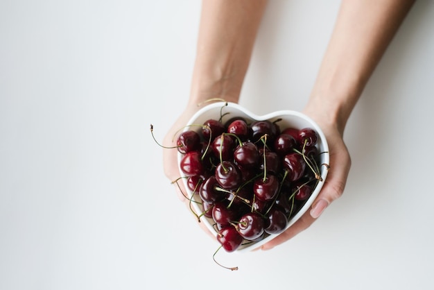 Cerises juteuses mûres dans une assiette en forme de coeur dans les mains des femmes sur fond blanc pour les restaurants et les menus avec espace de copie