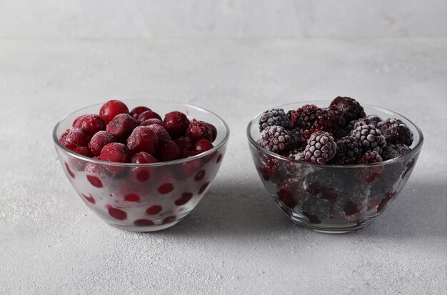 Cerises et mûres congelées dans deux bols transparents en verre sur fond de béton gris