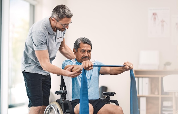 Cet exercice est également idéal pour maintenir un bon équilibre Photo d'un homme âgé en fauteuil roulant faisant de l'exercice avec une bande de résistance aux côtés de son physiothérapeute