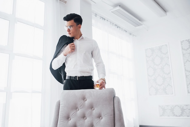 Avec chaise blanche devant. Un homme luxueux à l'usure classique se tient dans la pièce et tient le haut du costume et le verre avec de l'alcool.