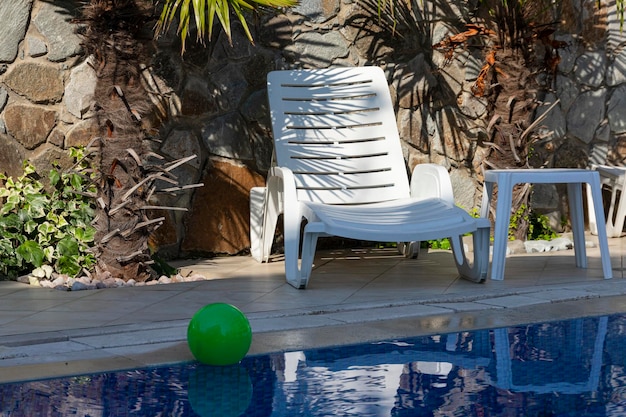 Chaise longue blanche au bord de la piscine avec de l'eau bleue et une boule verte sur fond de palmiers