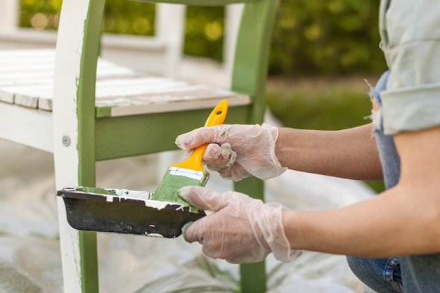 Chaise de peinture à la couleur verte avec une brosse dans des gants de protection Le travailleur peint les meubles de jardin en vert