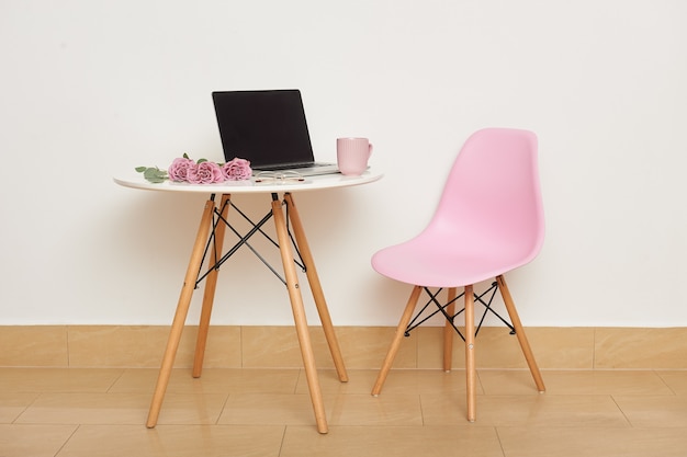 Une chaise rose et une table blanche contre le mur. Sur la table se trouvent un ordinateur portable, des lunettes, une tasse et un bouquet de roses. Intérieur de studio ou lieu de travail.