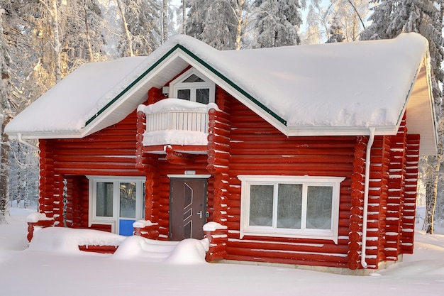 Chalet en bois de rondins peints en rouge, avec toit couvert de neige sur fond de forêt d'hiver enneigée dans la journée.