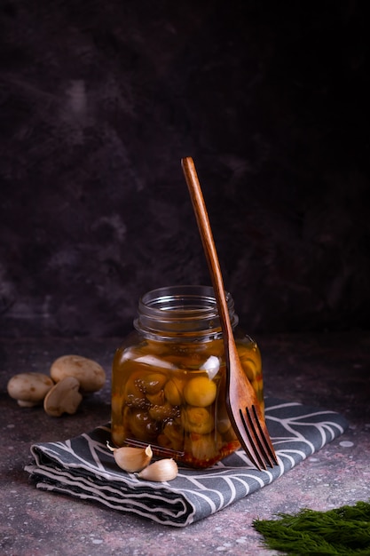 Champignons champignon en conserve fermentés avec une fourchette écologique en bois et dans un bocal en verre avec de l'ail, du laurier et de l'aneth sur une serviette grise