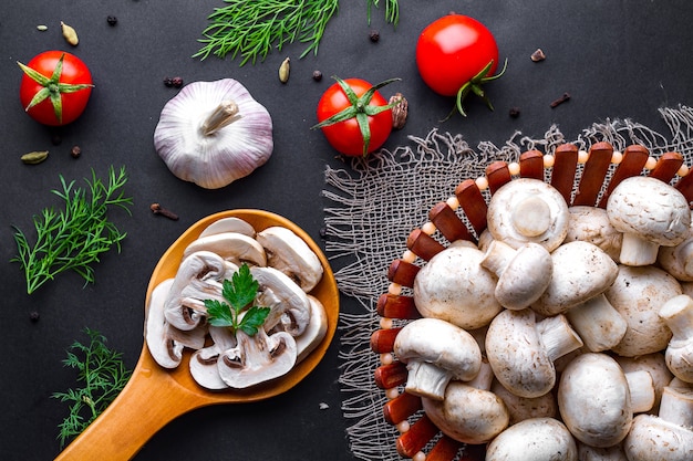 Champignons tranchés frais dans une cuillère en bois sur une surface noire. Cuire des plats maison de champignons blancs mûrs avec du persil, de l'aneth et des légumes.