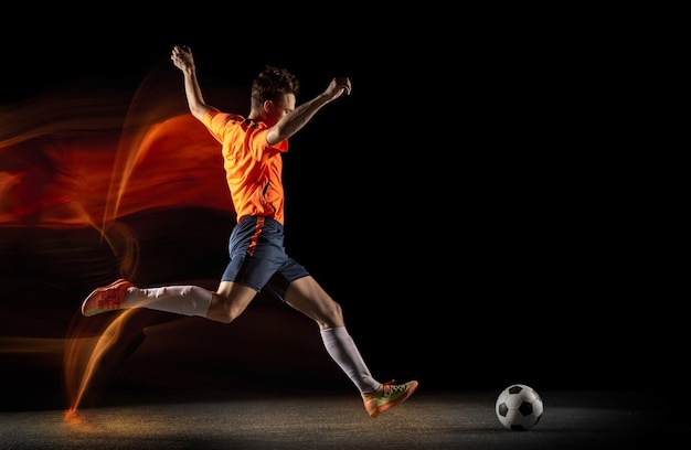 Champion. Jeune Joueur De Football Ou De Football Masculin Caucasien En Vêtements De Sport Et Bottes Frappant Le Ballon Pour Le But En Lumière Mixte Sur Fond Sombre. Concept De Mode De Vie Sain, Sport Professionnel, Passe-temps.