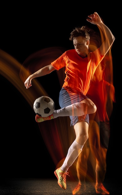 Champion. Jeune joueur de football ou de football masculin caucasien en vêtements de sport et bottes frappant le ballon pour le but en lumière mixte sur fond sombre. Concept de mode de vie sain, sport professionnel, passe-temps.