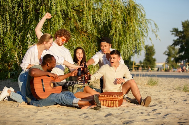 Chanson de jeunesse. Groupe d'amis trinquant à bière pendant le pique-nique à la plage au soleil. Mode de vie, amitié, s'amuser, week-end et concept de repos. Semble gai, heureux, célébrant, festiv