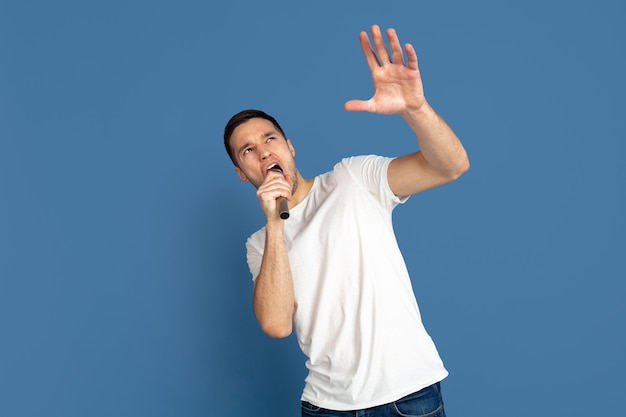 Chanter comme des célébrités, des stars. Portrait du jeune homme caucasien sur le mur bleu du studio.