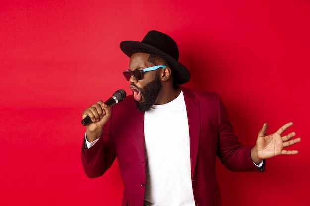 Chanteur noir passionné se produisant sur fond rouge, chantant dans un microphone, portant une tenue de fête, debout sur fond rouge.