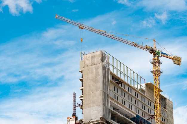 Chantier de construction avec grue et bâtiment. Secteur de l'immobilier. La grue utilise l'équipement de levage de bobine dans le chantier de construction. Bâtiment en acier et béton. Travail de grue contre le ciel bleu et les nuages blancs