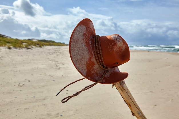 Un chapeau de cowboy orange saupoudré de sable est suspendu à un bâton noueux sur une plage déserte. Concept d'aventure