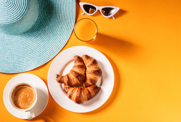Chapeau, croissant de lunettes de soleil avec une tasse de café et un verre de jus d'orange sur une table jaune au soleil