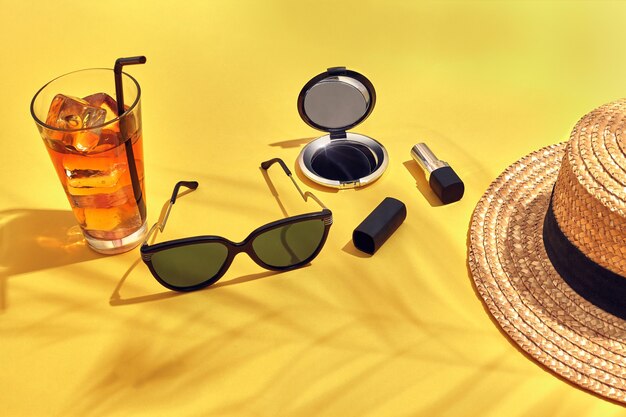 Chapeau de femme de plage de paille, boisson avec de la glace dans un verre et lunettes de soleil sur fond jaune