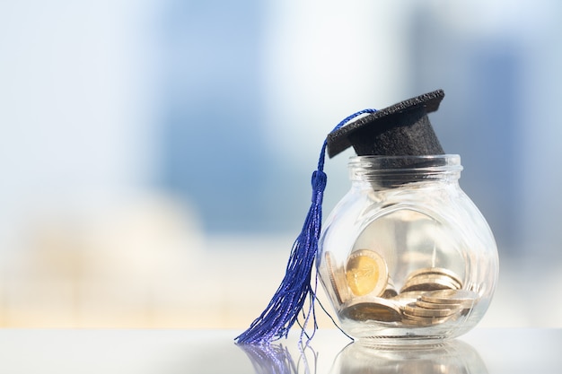 Chapeau de graduation avec gland bleu sur le bocal en verre rempli de pièces de monnaie sur fond de ville moderne