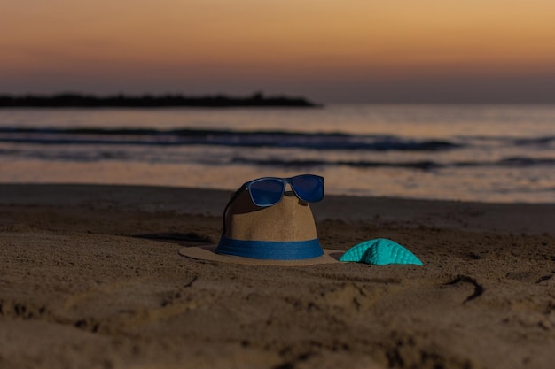 Chapeau avec des lunettes de soleil sur le dessus et une étoile de mer en plastique à côté placé sur le sable de la plage au lever du soleil concept de plage et d'été