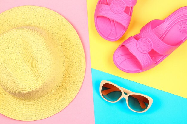 Chapeau, lunettes de soleil et pantoufles sur un fond pastel tricolore de bleu, jaune et rose.