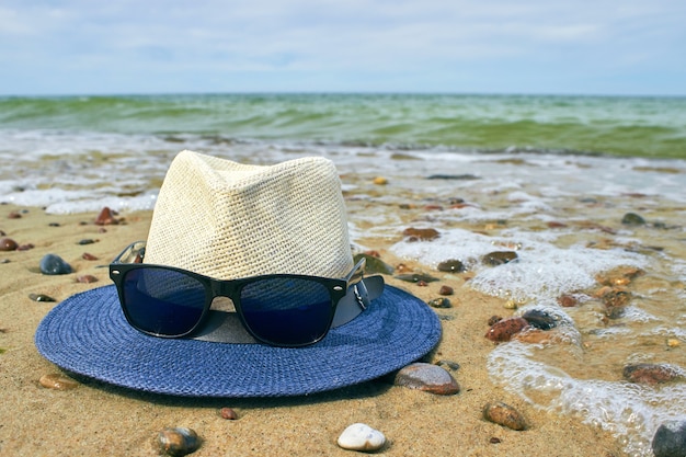 Chapeau de paille et lunettes de soleil se trouvent sur un bord de mer de sable avec des galets.