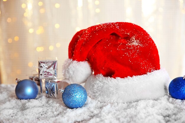 Chapeau de père Noël avec des boules et des coffrets cadeaux sur une table enneigée sur un mur de paillettes