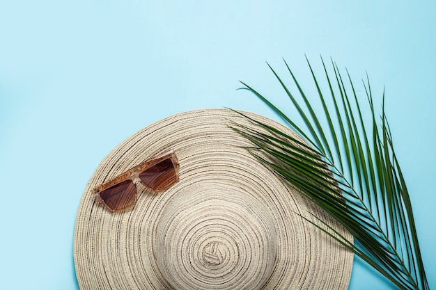 Chapeau pour femme à large bord, lunettes de soleil et branche de palmier sur fond bleu.