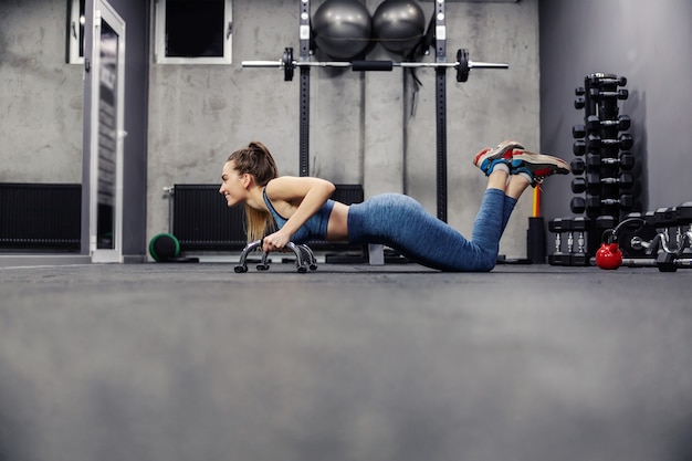 Chargez les pompes et la position de la planche. Vue latérale d'une jolie femme mince en tenue de sport et en bonne forme faisant des exercices de bras dans une salle de sport intérieure. Stabilité du corps, souplesse musculaire et vie sportive