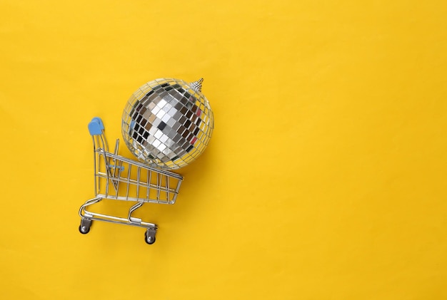 Chariot de magasinage avec boule disco sur fond jaune Concept de fête minimalisme