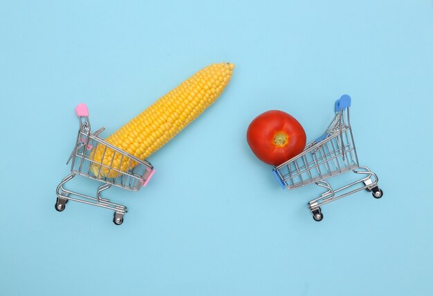 Chariots de supermarché avec balançoire de maïs et tomate sur fond bleu.