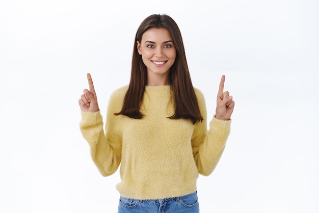 Charismatique confiante jeune femme amicale en chandail jaune demandant de tourner votre attention vers le haut la publicité recommande de cliquer sur la bannière pointant les doigts vers le haut et souriant