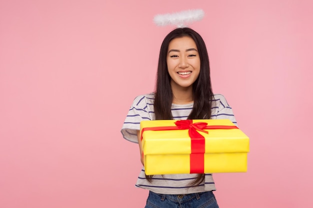 Charité et dons Portrait d'une fille asiatique angélique avec halo sur la tête donnant une boîte-cadeau à la caméra et souriant modestement exprimant la gentillesse générosité soins studio tourné isolé sur fond rose