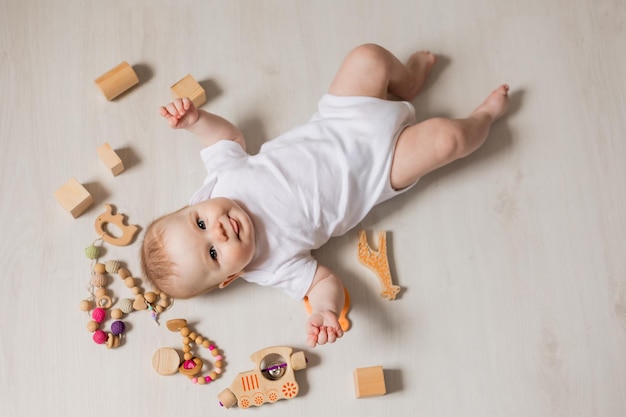 Charmant bébé en body blanc allongé sur le dos par terre parmi des hochets et des jouets éducatifs en bois. vue de dessus. Photo de haute qualité