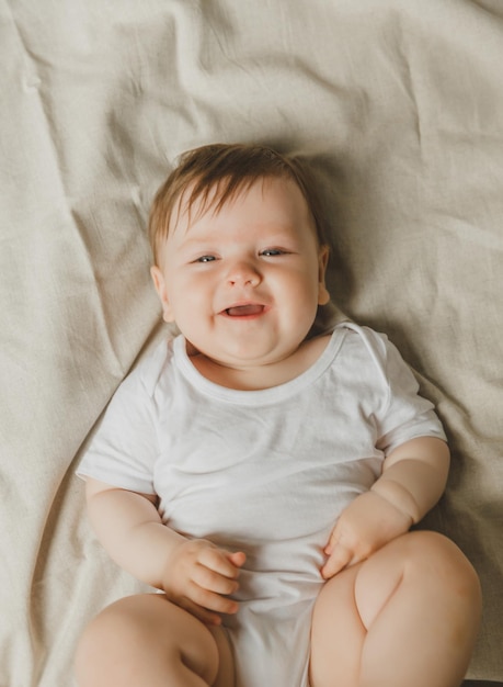 Un charmant bébé de six mois aux yeux bleus est allongé dans un lit dans un costume blanc.