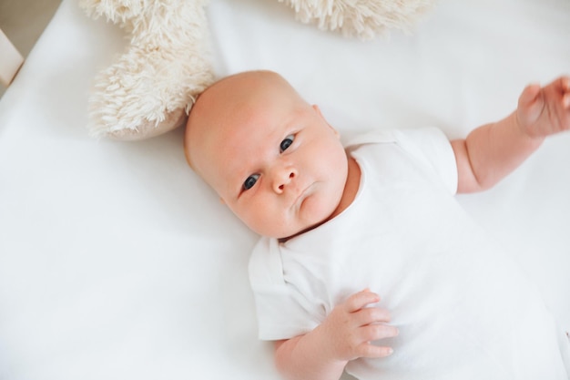 Un charmant bébé souriant de 2 mois aux yeux bleus dans un body blanc se trouve dans un berceau à côté d'un ours en peluche Vue de dessus un nouveau-né