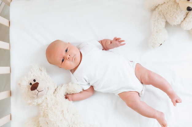 Un charmant bébé souriant de 2 mois aux yeux bleus dans un body blanc se trouve dans un berceau à côté d'un ours en peluche Vue de dessus un nouveau-né