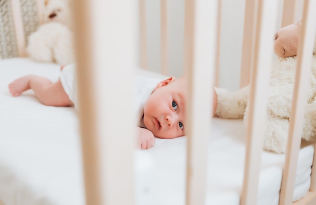 Un charmant bébé souriant de 2 mois aux yeux bleus dans un body blanc se trouve dans un berceau à côté d'un ours en peluche Vue de dessus un nouveau-né