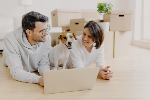 Un charmant couple juste marié allongé sur le sol utilise un ordinateur portable crée un projet de conception pour une nouvelle maison leur chien préféré pose entre eux un tas de boîtes en carton en arrière-plan Déménagement vers une nouvelle maison