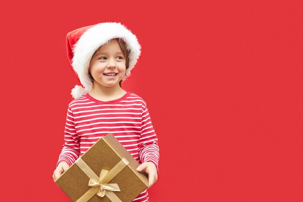 Charmant garçon avec un chapeau de Père Noël et une boîte à cadeaux dans les mains sur un fond rouge avec de l'espace pour le texte