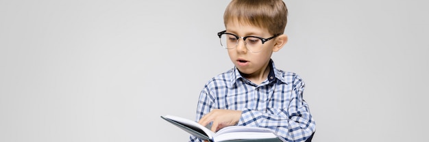 Un charmant garçon avec une chemise incrustée et un jean léger se dresse. Le garçon tient un livre dans ses mains. Garçon à lunettes