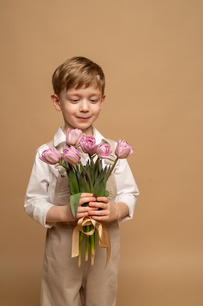 un charmant garçon de quatre ans en salopette beige et une chemise blanche tient un bouquet de tulipes roses