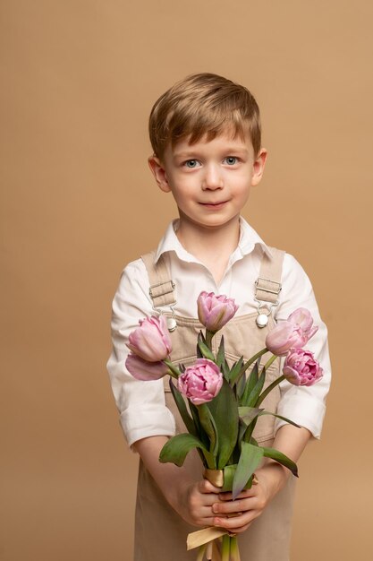 un charmant garçon de quatre ans en salopette beige et une chemise blanche tient un bouquet de tulipes roses