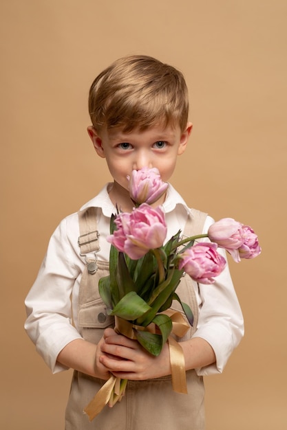 un charmant garçon de quatre ans en salopette beige et une chemise blanche tient un bouquet de tulipes roses