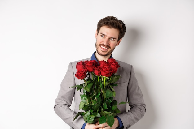 Charmant homme souriant passe rendez-vous romantique, vêtu d'un costume, tenant un bouquet de roses et regardant à gauche, debout le jour de la Saint-Valentin avec un cadeau sur fond blanc