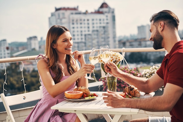 Charmant jeune couple en vêtements décontractés se portant un toast et souriant assis sur la terrasse sur le toit à l'extérieur