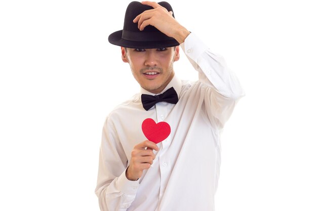 Charmant jeune homme aux cheveux noirs en T-shirt blanc avec noeud papillon et chapeau noir tenant un coeur en papier rouge sur fond blanc en studio.