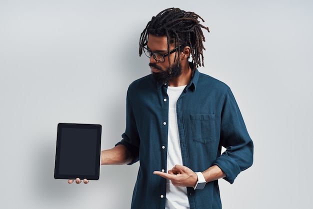 Charmant jeune homme en lunettes pointant vers l'espace de copie à l'aide d'une tablette numérique tout en se tenant contre un mur gris