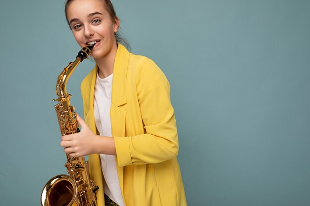 Charmante adolescente brune souriante et souriante portant une veste jaune à la mode, isolée sur un mur de fond bleu jouant du saxophone en regardant la caméra. Espace de copie