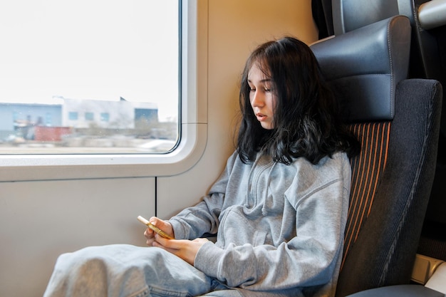 Charmante adolescente pensive assise à l'intérieur du chariot de train électrique de banlieue et utilisant un téléphone intelligent