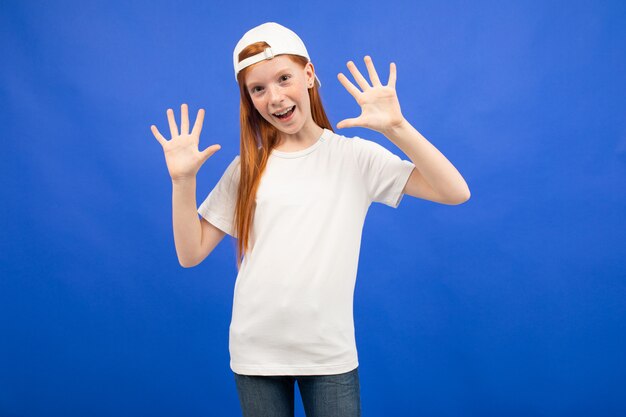 Charmante adolescente rousse dans un T-shirt blanc montre un espace d'impression vierge sur un studio bleu